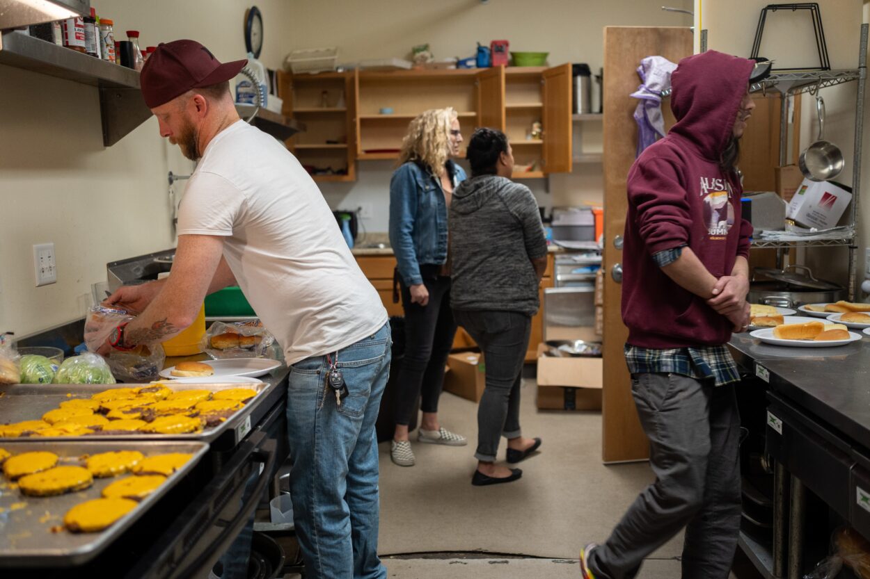 people stand in a kitchen, assembling cheese burgers and other food
