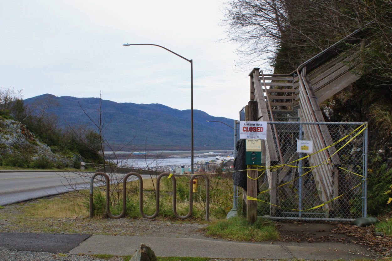 a closed sign in front of a fenced off entrance to a wooden staircase, ascending up into a hillside. a view of a channel and mountains in the background