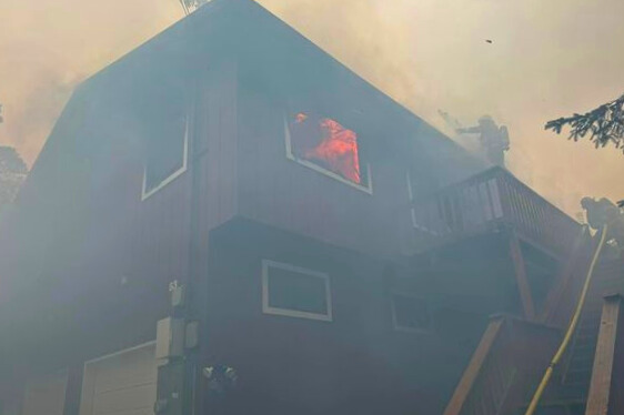 a smoke covered building with flames coming from one window, a firefighter pulling a yellow hose up a flight of stairs and another apparently hosing down the roof